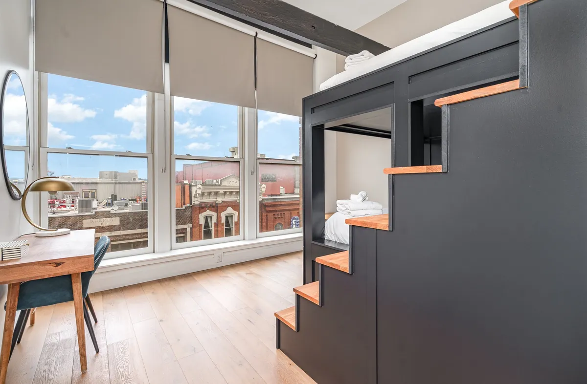 A modern lofted sleeping and working space with a bed on a raised platform accessed by wooden stairs, large windows offering city views, and a minimalist desk and chair set up for productive work. The space feels airy, functional, and welcoming.