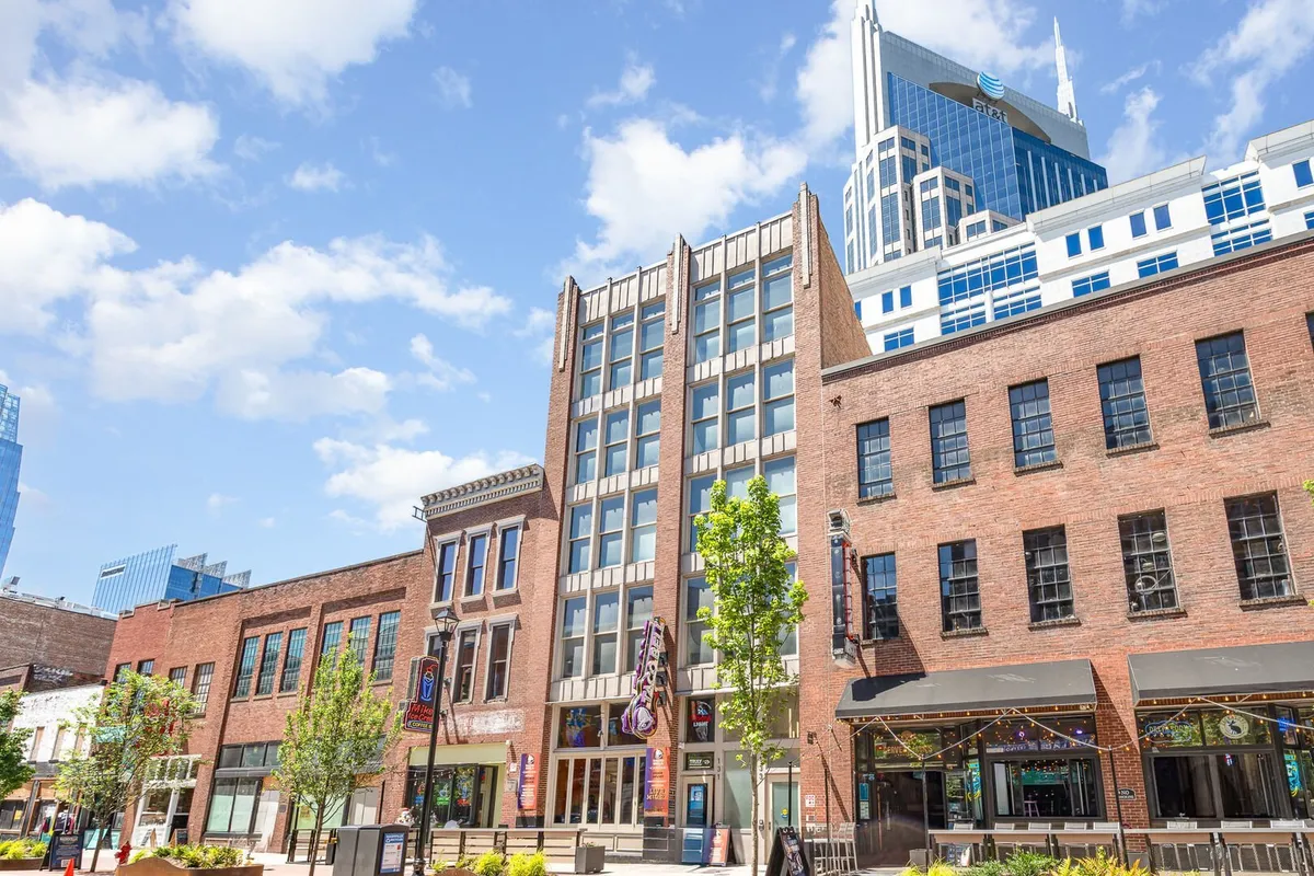 A lively street scene with historic brick buildings, modern high-rises in the background, and lush greenery in planters along the sidewalk. Outdoor cafes and people exploring boutiques add to the energetic, walkable city atmosphere.