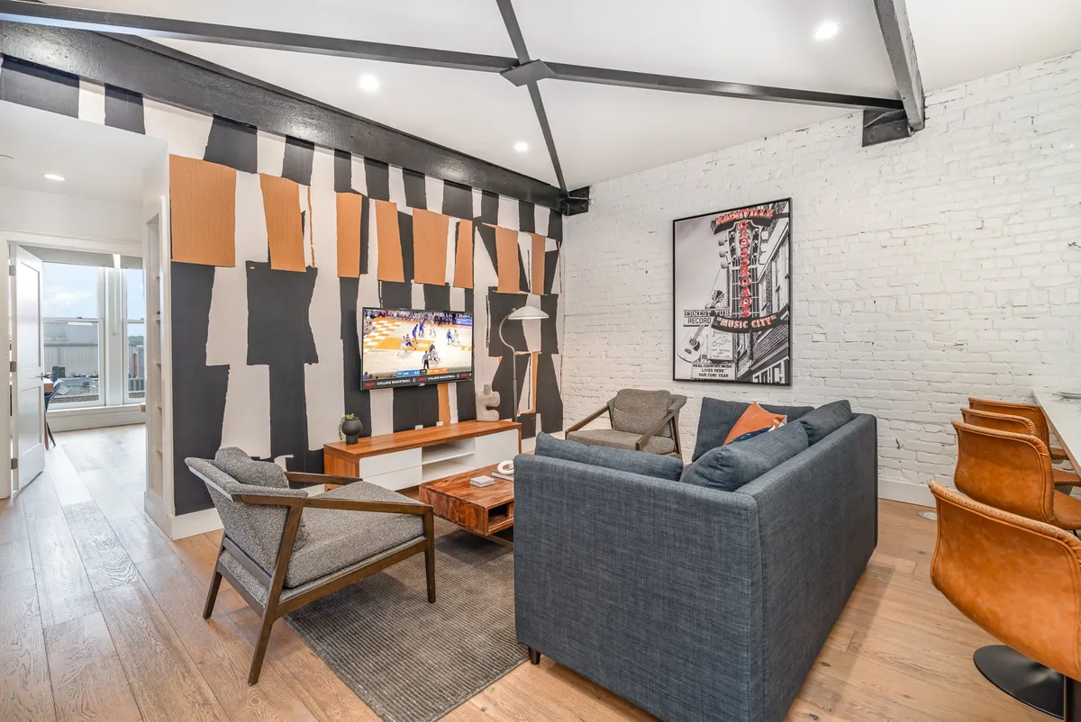 A spacious living area featuring a large TV mounted on the wall, modern artful accents, a comfortable sectional seating arrangement, and a wooden coffee table in the center. The space is bright and airy, ideal for lounging or hosting group activities. The Smart TV and ample seating perfectly support entertainment and socialization.