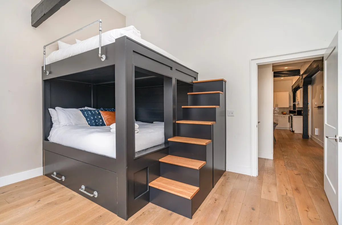 The image displays a stylish bunk bed setup featuring king-sized bedding, a built-in staircase for easy access to the top bunk, crisp linens, and playful decorative pillows. The overall mood is warm and inviting, suggesting both fun and comfort for groups or families sharing the space.