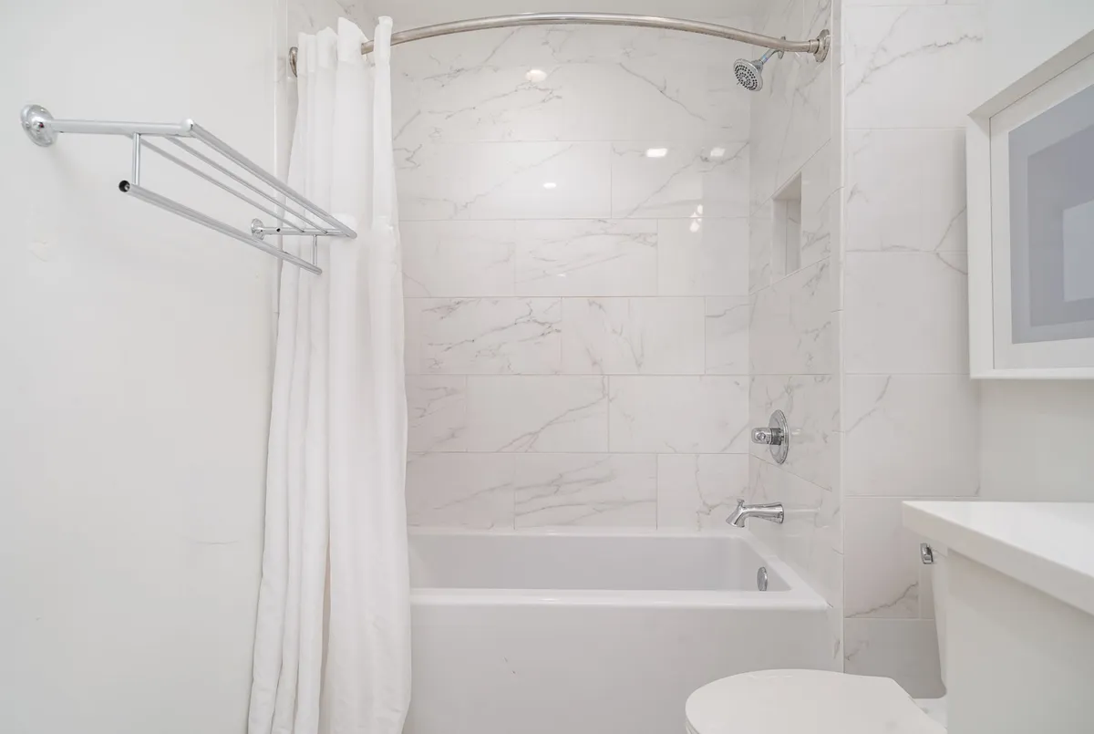 A serene bathroom with a white bathtub and shower combo, a modern showerhead, marble-tiled walls, a white shower curtain, and a sleek sink. The space is bright and looks clean, perfect for relaxing or getting ready.