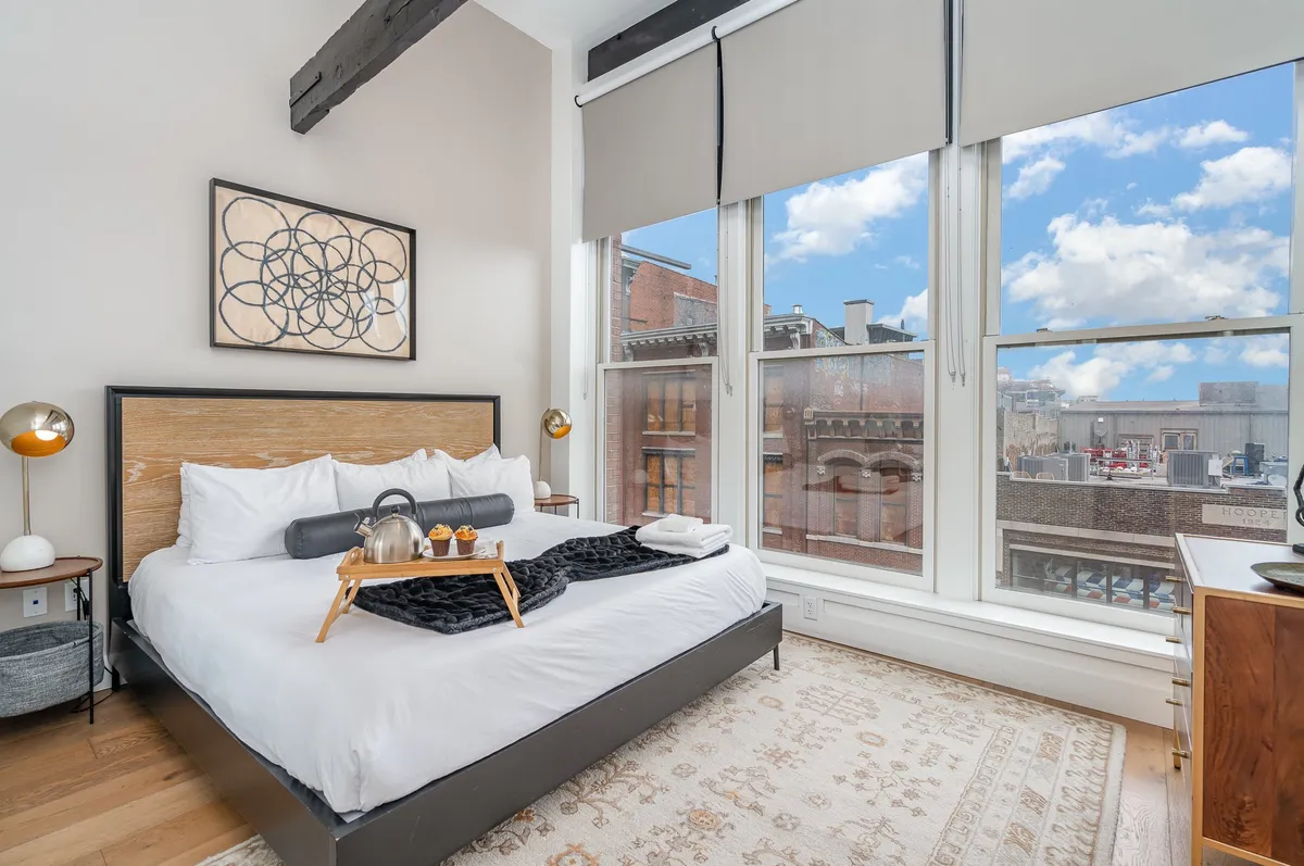 A sunlit bedroom with a plush king bed adorned in white linens, large windows letting in natural light, and tasteful artwork above the headboard. A breakfast tray atop the bed suggests relaxation and a leisurely morning routine, reinforcing a sense of luxury and calm.