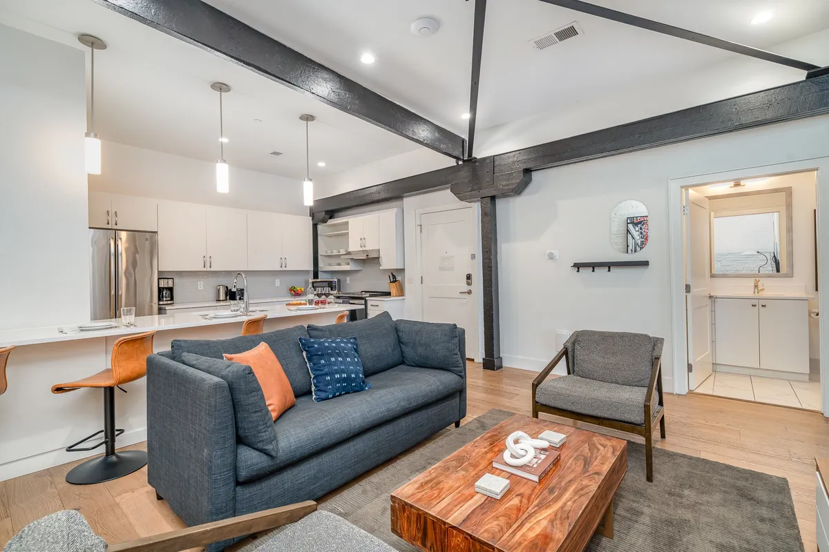 A cozy and modern living area with a plush sofa decorated with colorful cushions, a sturdy wooden coffee table, and an adjacent open kitchen space. The layout invites conversation, shared snacks, and group relaxation, echoing the twin lounge concept.