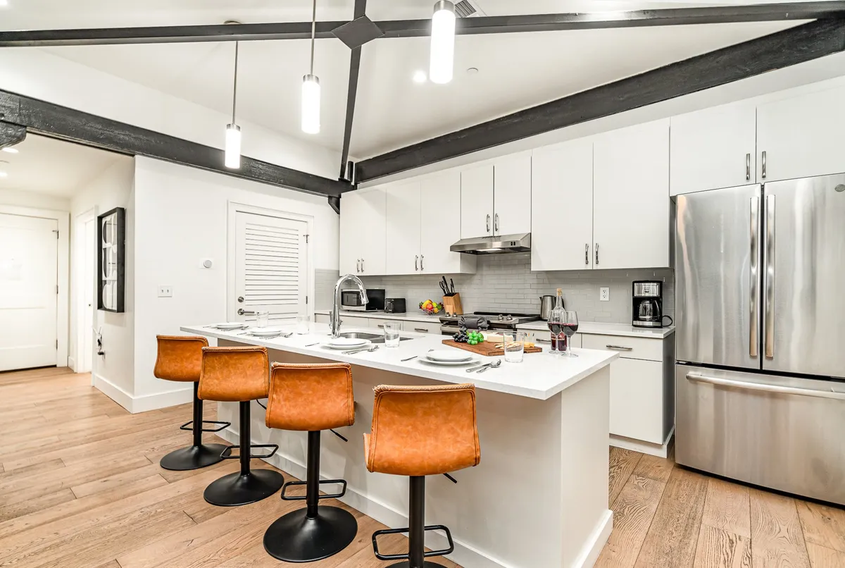 A modern kitchen featuring an island counter with bar stools, stainless steel appliances, open shelving displaying kitchenware, and touches of decorative fruits and plants. The space is open and inviting, perfect for both cooking and socializing.
