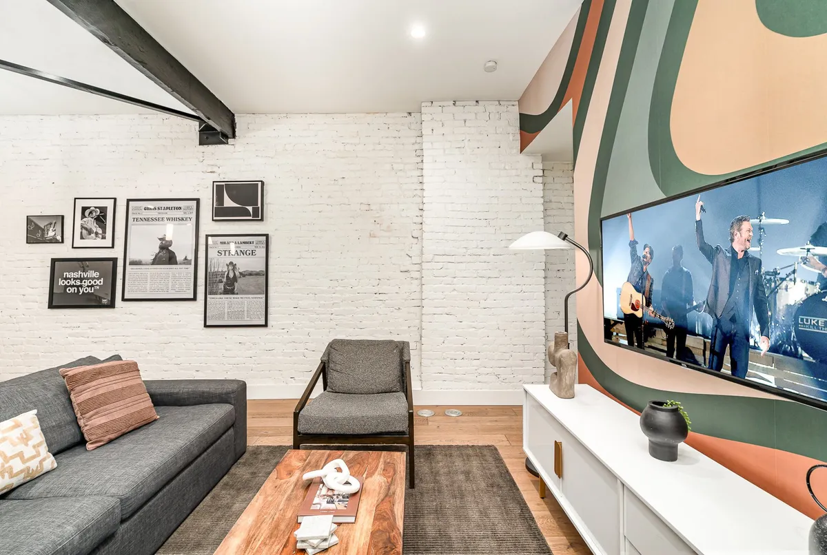 A comfortable living room setup with a sofa, large TV displaying a performance scene, music-themed wall art, and a wooden coffee table. The space is designed for both entertainment and relaxation, with decor that hints at music and social gatherings.