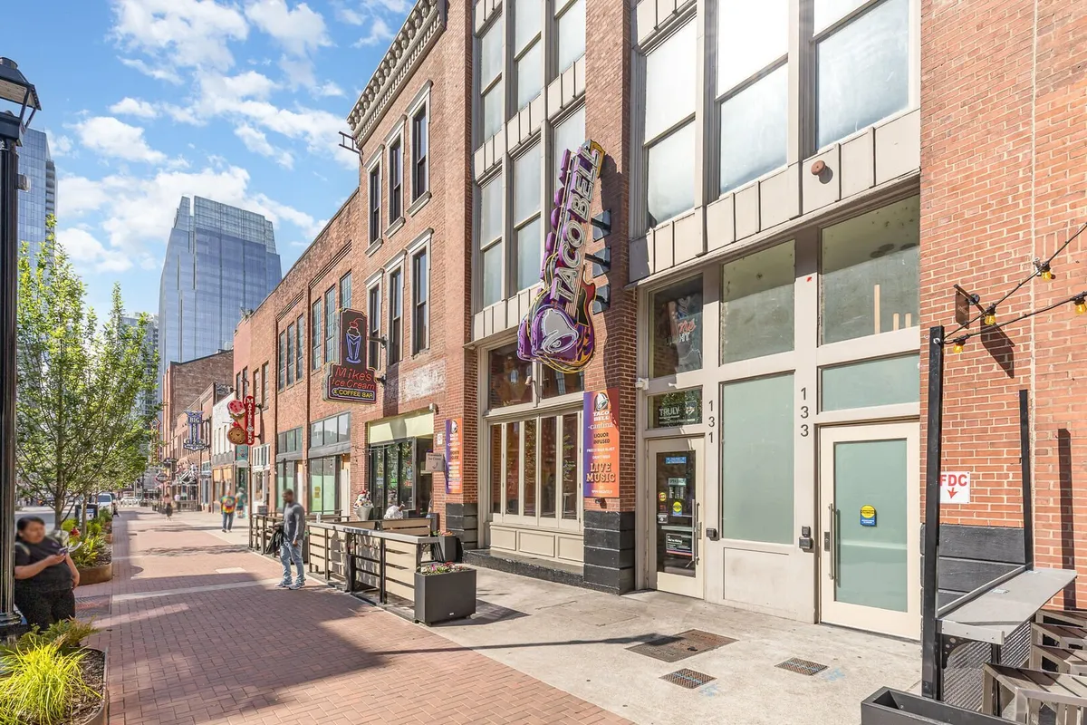 A vibrant downtown street scene featuring colorful storefronts, brick architecture, and energetic sidewalk seating. Diners enjoy meals at outdoor cafes while people stroll and shop, capturing the lively spirit of the area.